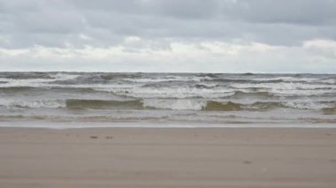 Letonya 'daki Baltık kıyılarının boş kumsallarına deniz dalgaları çarpıyor. Jurmala kıyı şeridinde bulutlu ve köpüklü su. Soğuk sonbahar ya da kış gününde deniz gelgiti olan kıyı manzarası.