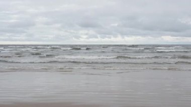 Deniz dalgaları bulutlu bir gökyüzünün altındaki geniş kumlu bir plaja yuvarlanıyor. Ön planda ıslak kum dalgaları olan Baltık deniz kıyısı manzarası. Sonbahar mevsiminde deniz kenarında kasvetli rüzgarlı hava.