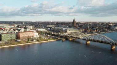 Riga Demiryolu Köprüsü ile Daugava nehri üzerinde hareket eden tren arasında Letonya 'nın kuzey Avrupa şehrinde güneşli bir günde Riga Merkez Market hangarları ve Letonya Bilim Akademisi gökdelenleri yer alıyor..
