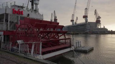 HELSINKI, FINLAND - 12 Aralık 2025 Hietalahti limanındaki bir restoran teknesinin kırmızı kürekli çarkı. Endüstriyel tersane vincleri bulutlu bir kış gökyüzünün altında duruyor..