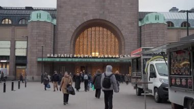 HELSINKI, FINLAND - 10 Aralık 2025: Yayalar Helsinki Merkez Tren İstasyonu 'nun aydınlık ana girişine doğru yürürler sokak yemek kamyonlarını geçerek. Tarihi granit cephe ve şehir merkezi.