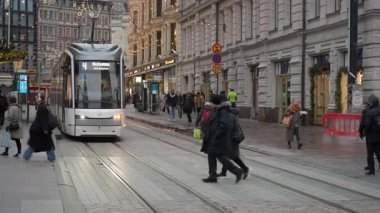 HELSINKI, FINLAND - 12 Aralık 2025: Aleksanterinkatu boyunca giden modern bir Skoda tramvay hattı. İnsanlar akşam karanlığında Noel ışıklarının altında raylardan geçiyorlar..