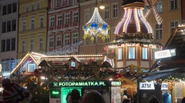 Gece Wroclaw Noel Pazarı 'nda ATM tabelaları ve fotoğraf standı olan şenlik tezgahları. Polonya 'da renkli yüzleri ve parlayan ışıklarıyla tarihi binaların yakınlarındaki tatil fuarını ziyaret eden kalabalık görülüyor.