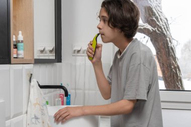 Gri tişörtlü çocuk banyo aynasında sakal tıraşı yapmak için elektrikli tıraş makinesi kullanıyor. Sabah rutini, erkek derisi, ergenlik çağı ve ilk tıraş prosedürü sağlık konsepti..