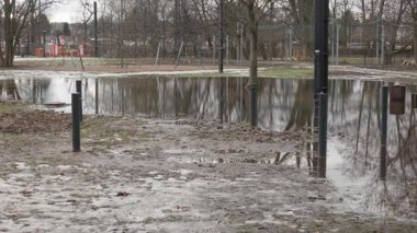 Dağınık bahar buzları çözülürken şehir parkındaki oyun alanı ekipmanlarını yansıtan büyük bir su birikintisi. Şehir meydanındaki ıslak çamurlu zeminde kar ve buz eridi. Kasvetli hava ve mevsimsel geçiş havası.