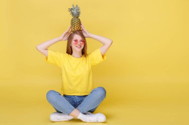 Pembe gözlüklü duygusal kız sarı arka planda ananasla oturuyor.