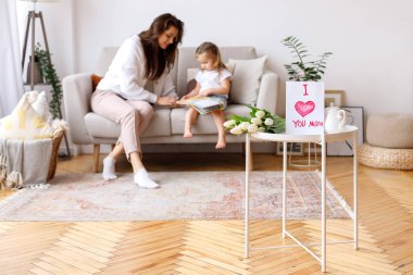 Oturma odasındaki Uluslararası Kadınlar Günü kartpostalı anne ve kızının arka planında.