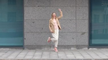 Gri duvarın yanında dans eden ve gülümseyen sarışın kız.