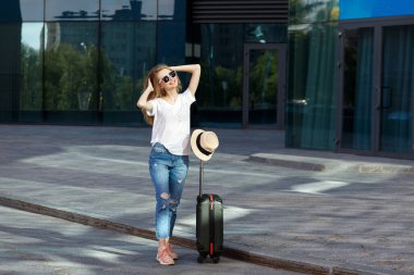 Yazın güneş ışığında tekerlekli bavullu, gözlüklü sarışın. seyahat ve tatil