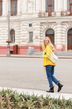 Sarı yağmurluklu ve lastik çizmeli bir kadın sonbaharda şehirde yürüyüş yapıyor.