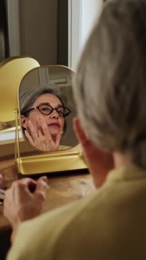 Gri saçlı zarif olgun bir kadın aynaya bakarken nemlendirici sürüyor. Günlük cilt bakımı sağlıklı, parlayan cilt için