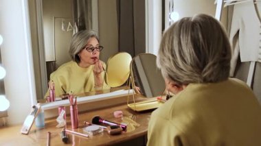 Şık, yaşlı bir kadın soyunma odasında ruj sürüyor ve kameraya gülümsüyor. Günlük güzellik rutini ve olgun cilt bakımı