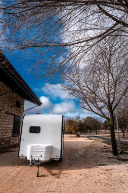 Taştan bir binanın yanındaki çakılların üzerine park edilmiş beyaz karavan ve mavi gökyüzünün altında çıplak ağaçlar. Kış kampı için ideal.