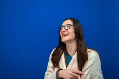 Uzun siyah saçlı ve gözlüklü bir kadın gülüyor, neşe ve iyimserliğini dile getiriyor. Mavi arka planda beyaz bir ceket giyiyor.