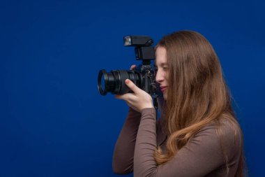 Genç fotoğrafçı kadın fotoğraf çekiyor, elinde mavi arka planda flaşlı profesyonel bir dijital kamera var.