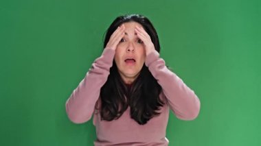 Neşeli esmer kadın komik suratlar yapıyor ve elleri yeşil perde arkasında kalp şekli oluşturuyor.