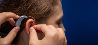 Elleri modern bej ve gri işitme cihazını bir kulağın arkasına sığdırarak, daha iyi duyma ve erişilebilirliği teşvik eder.