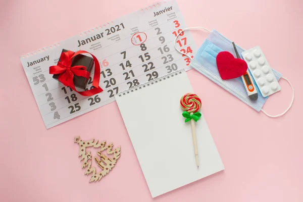 Flat lay on a pink background lies a notebook with a lollipop handle, a sheet of calendar 2021, a medical mask, a thermometer, a heart and a tablet. — Fotografia de Stock