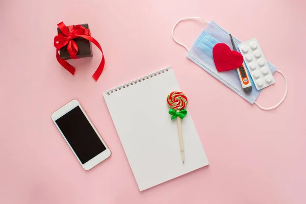 Piatto giaceva il giorno di San Valentino su uno sfondo rosa si trova un taccuino con una penna, uno smartphone, regalo, una maschera medica, un cuore, pillole e un termometro. — Foto Stock