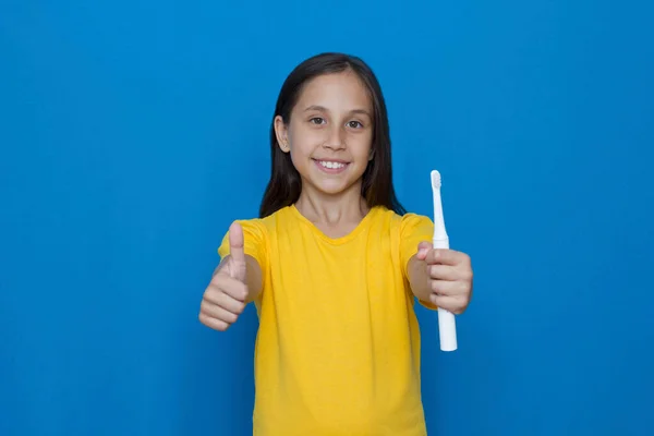 Brunett flicka med långt hår i en gul T-shirt på en blå bakgrund med en elektrisk tandborste visar cool, klass, super. — Stockfoto
