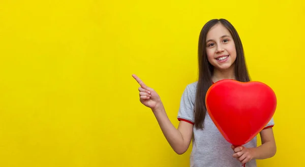Banner. Uma menina sorridente com cabelos longos em um fundo amarelo segura uma bola na forma de um coração. — Fotografia de Stock
