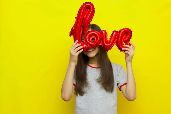 Uma menina sorridente com longos cabelos escuros em um fundo amarelo segura uma bola com a inscrição amor, espreita. — Fotografia de Stock