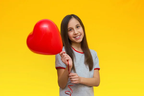 Uma menina sorridente e positiva com longos cabelos escuros em uma camiseta cinza em um fundo amarelo segura um coração de balão. — Fotografia de Stock