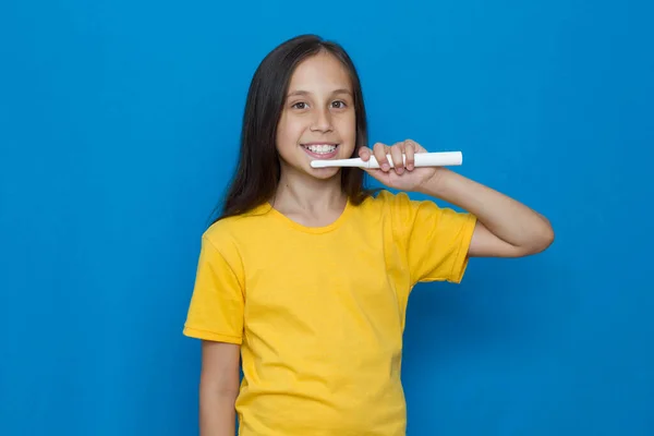 Brunett flicka med långt hår i en gul T-shirt på en blå bakgrund borstar tänderna med en elektrisk tandborste. — Stockfoto