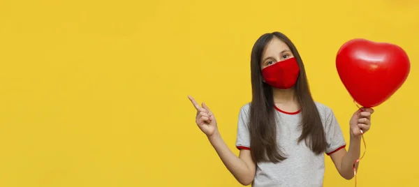 Banner. Menina com cabelos longos e escuros Usar uma camiseta cinza e máscara médica protetora em um fundo amarelo segura um balão vermelho. — Fotografia de Stock