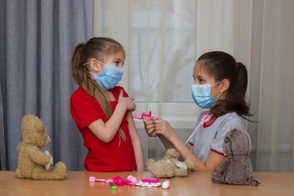 Δύο κορίτσια σχολικής και προσχολικής ηλικίας, φορώντας ιατρικές μάσκες, παίζουν στο νοσοκομείο με παιχνίδια, εμβολιάζονται. Εικόνα Αρχείου