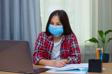 Tıbbi maskeli esmer bir kız, kırmızı gömlekli evde oturuyor, çalışıyor, ödevlerini yapıyor, internetten iletişim kuruyor..