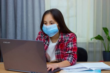 Tıbbi maskeli esmer bir kız, kırmızı gömlekli evde oturuyor, çalışıyor, ödevlerini yapıyor, internetten iletişim kuruyor..