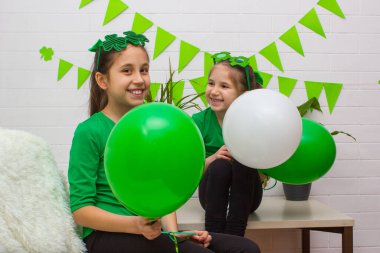 Yeşil tişört ve gözlüklü yonca giymiş iki kız kardeş ellerinde balonlarla Aziz Patrick Günü 'nü kutluyorlar..