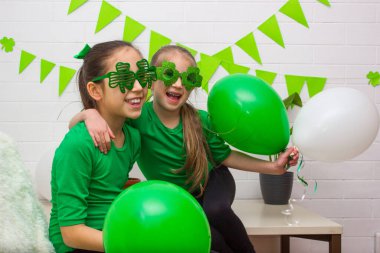 Yeşil tişört ve gözlüklü yonca giymiş iki kız kardeş ellerinde balonlarla Aziz Patrick Günü 'nü kutluyorlar..