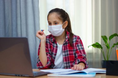 Tıbbi maskeli esmer bir kız, kırmızı gömlekli evde oturuyor, çalışıyor, ödev yazıyor, internetten iletişim kuruyor..