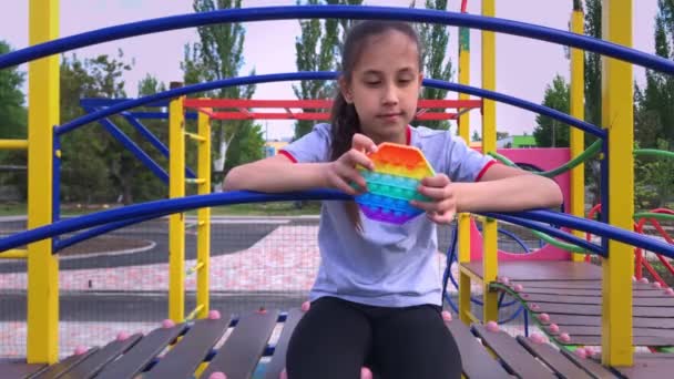A girl with dark hair in a striped T-shirt, upset, sad, sits on the slide of the playground, calming down, playing pop it, popit — Stok Video