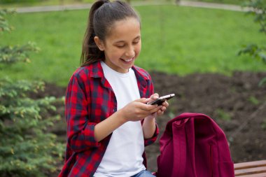 Uzun siyah saçlı, beyaz tişörtlü ve ekose gömlekli neşeli bir kız parkta oturuyor, gülüyor ve telefonla haberciyle iletişim kuruyor. Seçici odak.