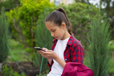 Beyaz tişörtlü, parktaki sırt çantalı, uzun siyah saçlı ve ekose gömlekli bir kız, elçiyle iletişim kurmak için telefonu hevesle kullanıyor. Seçici odak