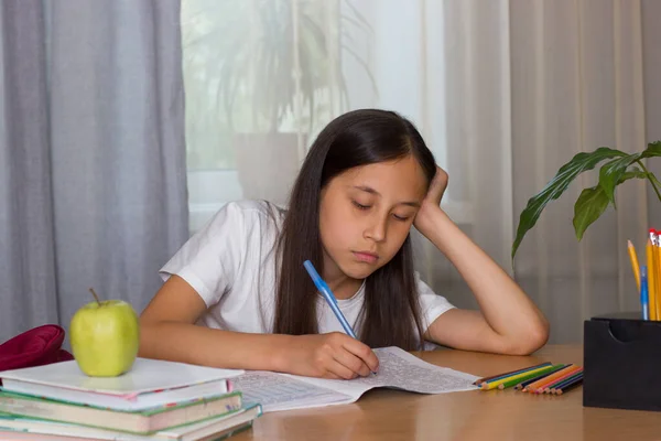 Une fille triste avec les cheveux longs foncés dans un T-shirt blanc s'assoit à la table à la maison, faisant ses devoirs, ses devoirs. Concept de retour à l'école. — Photo