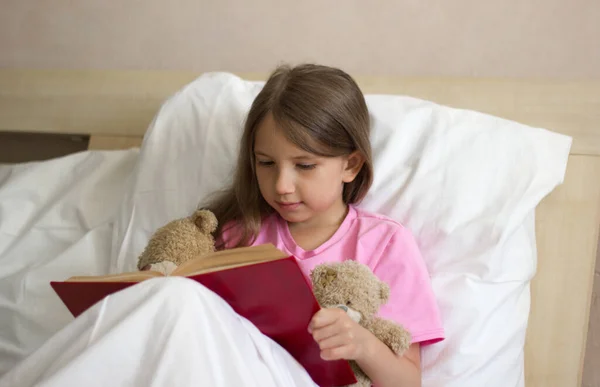 Ένα μικρό όμορφο κορίτσι με μακριά ξανθά μαλλιά με ροζ μπλουζάκι κάθεται στο κρεβάτι τη μέρα και διαβάζει ένα βιβλίο σε ένα αρκουδάκι. — Φωτογραφία Αρχείου