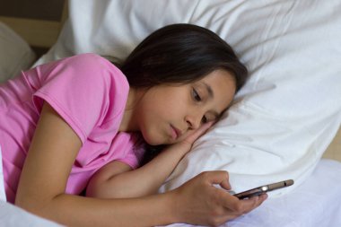 Uzun saçlı, pembe tişörtlü, üzgün bir esmer kız, yatağa uzanıyor ve elinde bir cep telefonu tutuyor.
