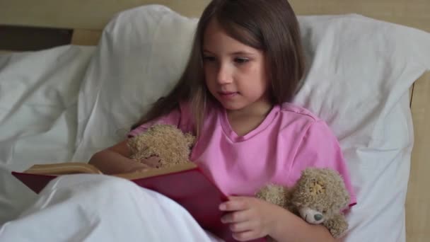 Una niña bonita con el pelo largo y rubio en una camiseta rosa se sienta en la cama durante el día y lee un libro a un oso de peluche — Vídeo de stock