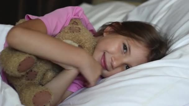 Egy csinos kislány, szőke hajjal, rózsaszín pólóban fekszik az ágyban, mosolyog és ölelget egy plüssmacit.