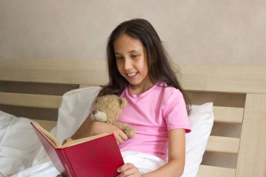 Pembe tişörtlü, uzun sarı saçlı güzel bir kız gündüzleri yatakta oturup oyuncak ayıya kitap okuyor.