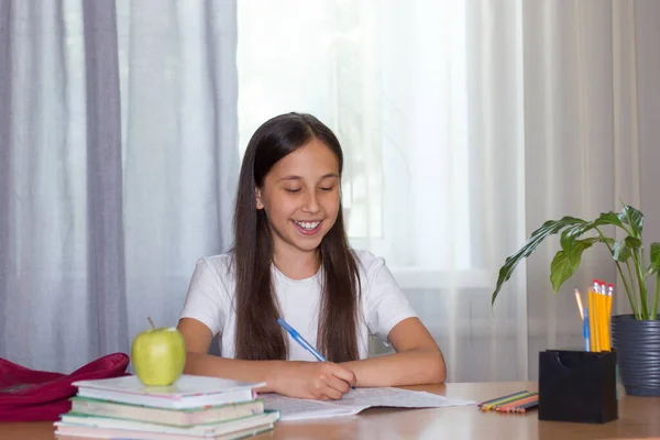 Ένα κορίτσι με σκούρα μακριά μαλλιά με λευκό μπλουζάκι κάθεται σε ένα τραπέζι, γράφει, διαβάζει. Επιστροφή στο σχολείο έννοια — Φωτογραφία Αρχείου