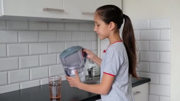 Uma morena em uma camiseta cinza na cozinha derrama água filtrada de um jarro em um copo. Conceito de estilo de vida saudável — Vídeo de Stock