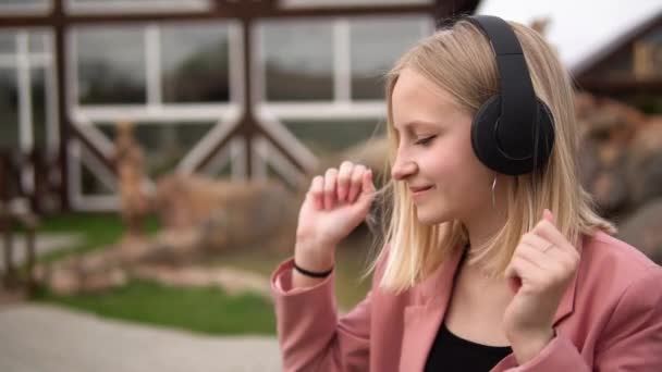 Gyönyörű fiatal lány szőke haj hallgat zenét fejhallgató, utcai stílus, szabadtéri portré. — Stock videók