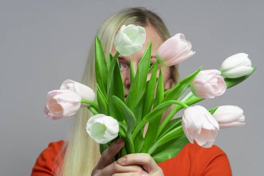 Kız önünde bir buket lale tutuyor, çiçekçi pastel arka planda, insan yüzünü bir buketin arkasına gizliyor.