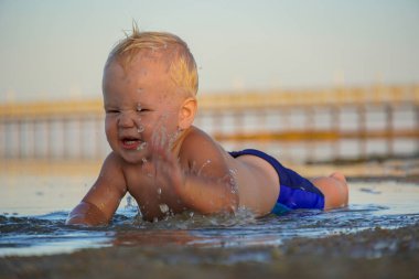 Sarı saçlı küçük bir çocuk kumsalda uzanıyor ve gülüyor..