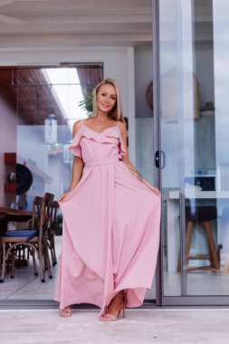 Lüks zengin villasında tatilde olan zarif, pembe uzun elbiseli beyaz kadın portresi.